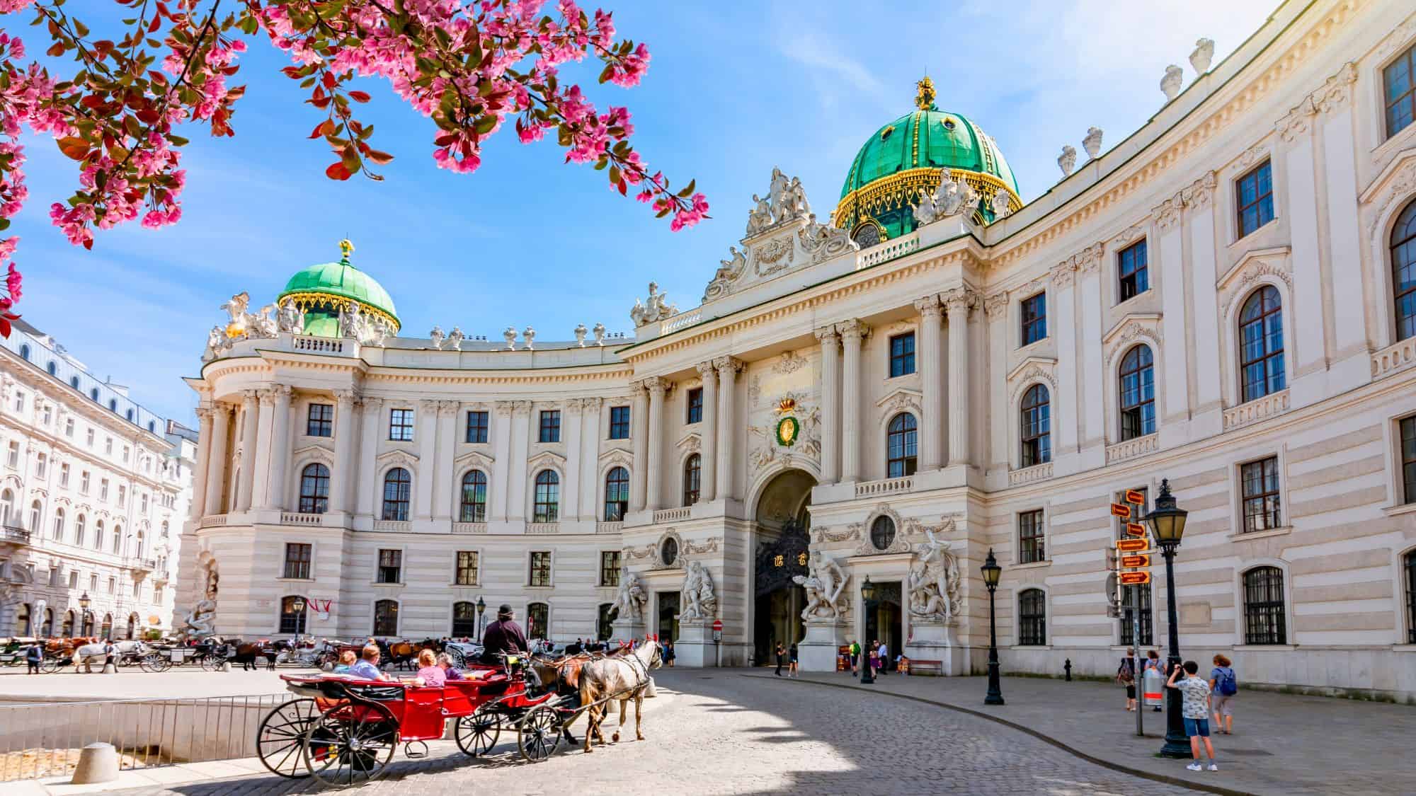 The curved façade of the Hofburg Palace, topped with a green dome, frames a bustling square with horse-drawn carriages.