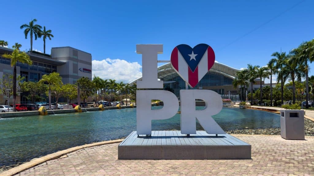 A large sculpture reading "I ❤️ PR" with the Puerto Rican flag in the heart stands by a reflecting pool outside the San Juan Convention Center on a sunny day.