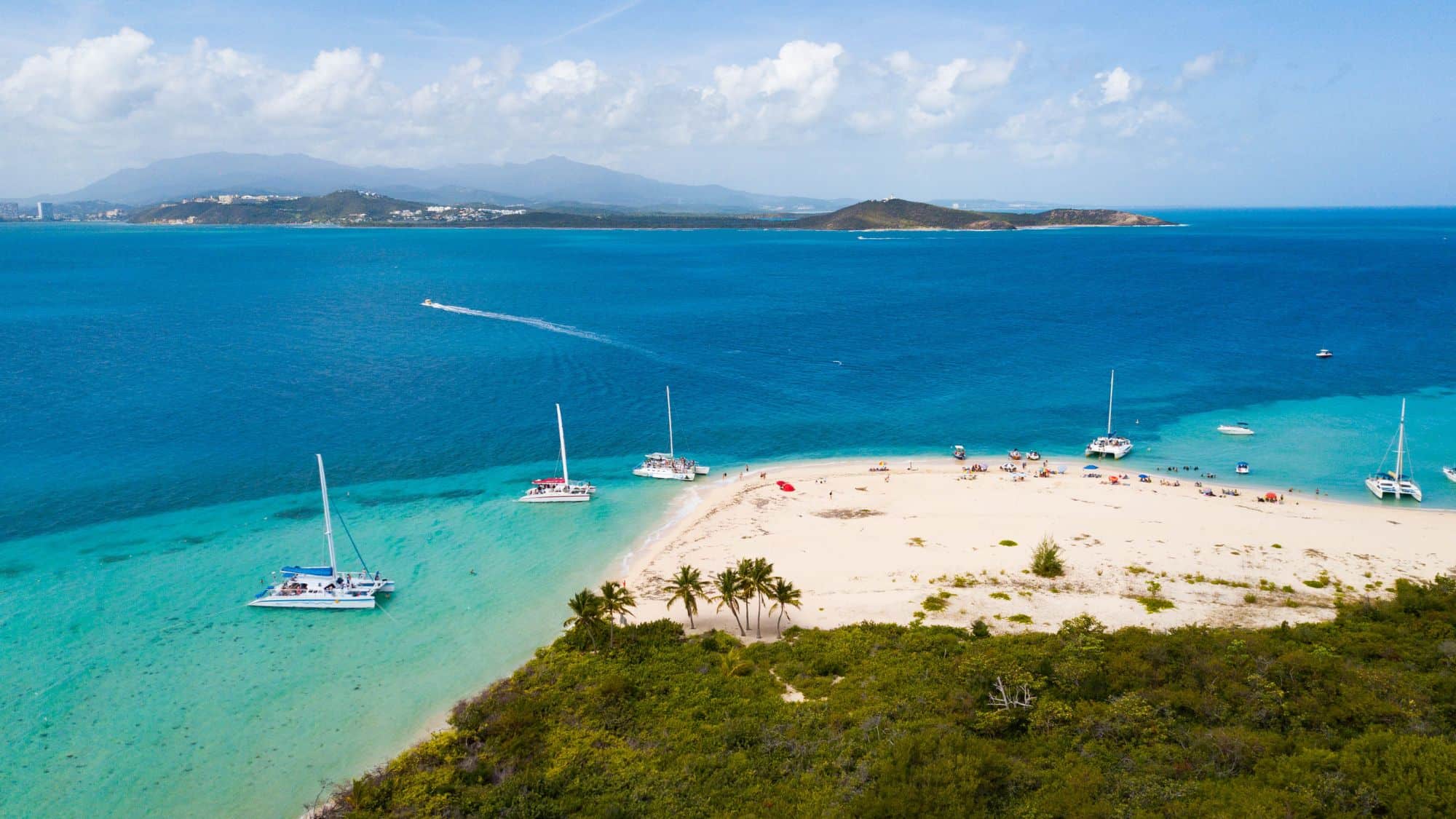 Catamarans are anchored in shallow turquoise waters beside a white sand beach on Icacos Island, with sunbathers relaxing and a view of the Puerto Rican mainland in the background.