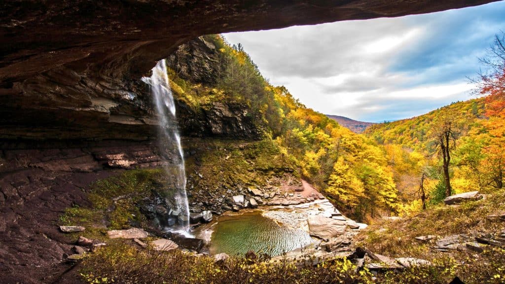 A tall waterfall cascades from a rocky overhang into a circular pool surrounded by trees glowing with fall colors in the Catskill Mountains.