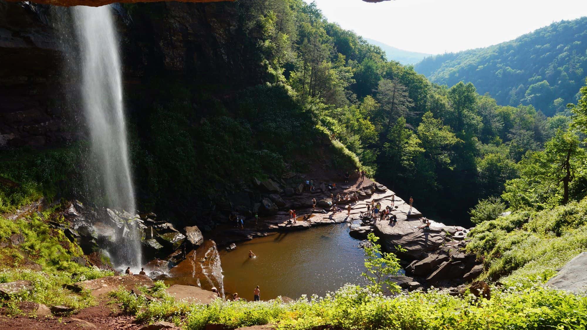 Visitors gather and swim around the base of Kaaterskill Falls, surrounded by lush green foliage and rocky cliffs under bright summer light.