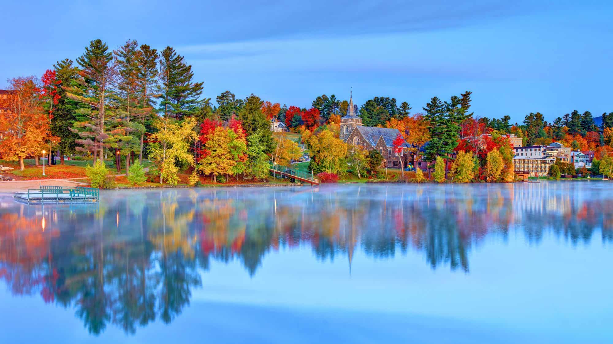 Brilliant fall foliage surrounds Lake Placid, with trees and a stone chapel mirrored perfectly in the calm, mist-tinted water.