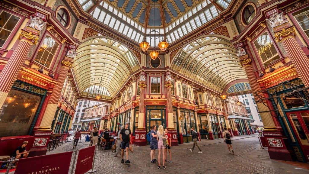 The ornate Victorian architecture of Leadenhall Market glows under a vaulted glass roof, with visitors walking between colorful shopfronts.