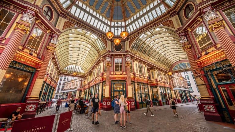 The ornate Victorian architecture of Leadenhall Market glows under a vaulted glass roof, with visitors walking between colorful shopfronts.