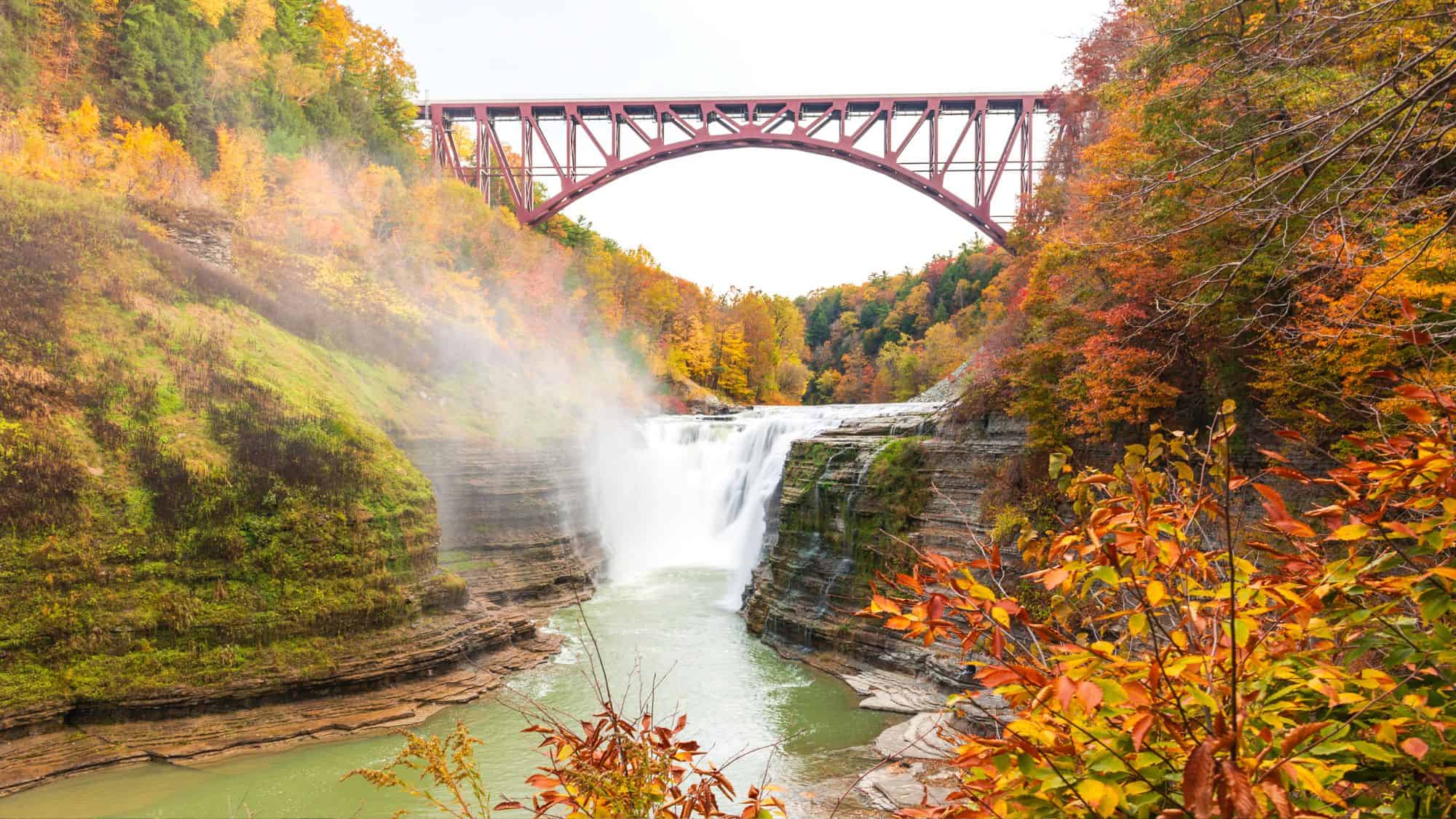 Middle Falls plunges into a rocky gorge framed by bright autumn leaves, with a striking red railway bridge spanning the canyon overhead.