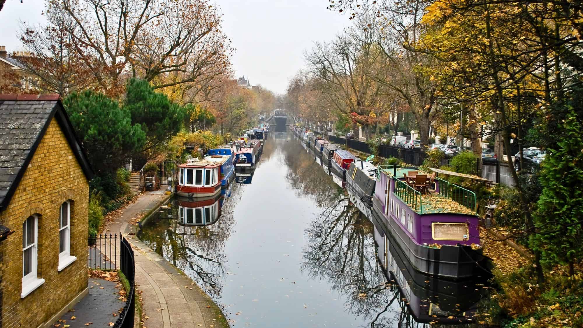 Narrowboats line a calm canal flanked by autumn trees, with reflections rippling under a soft gray sky.