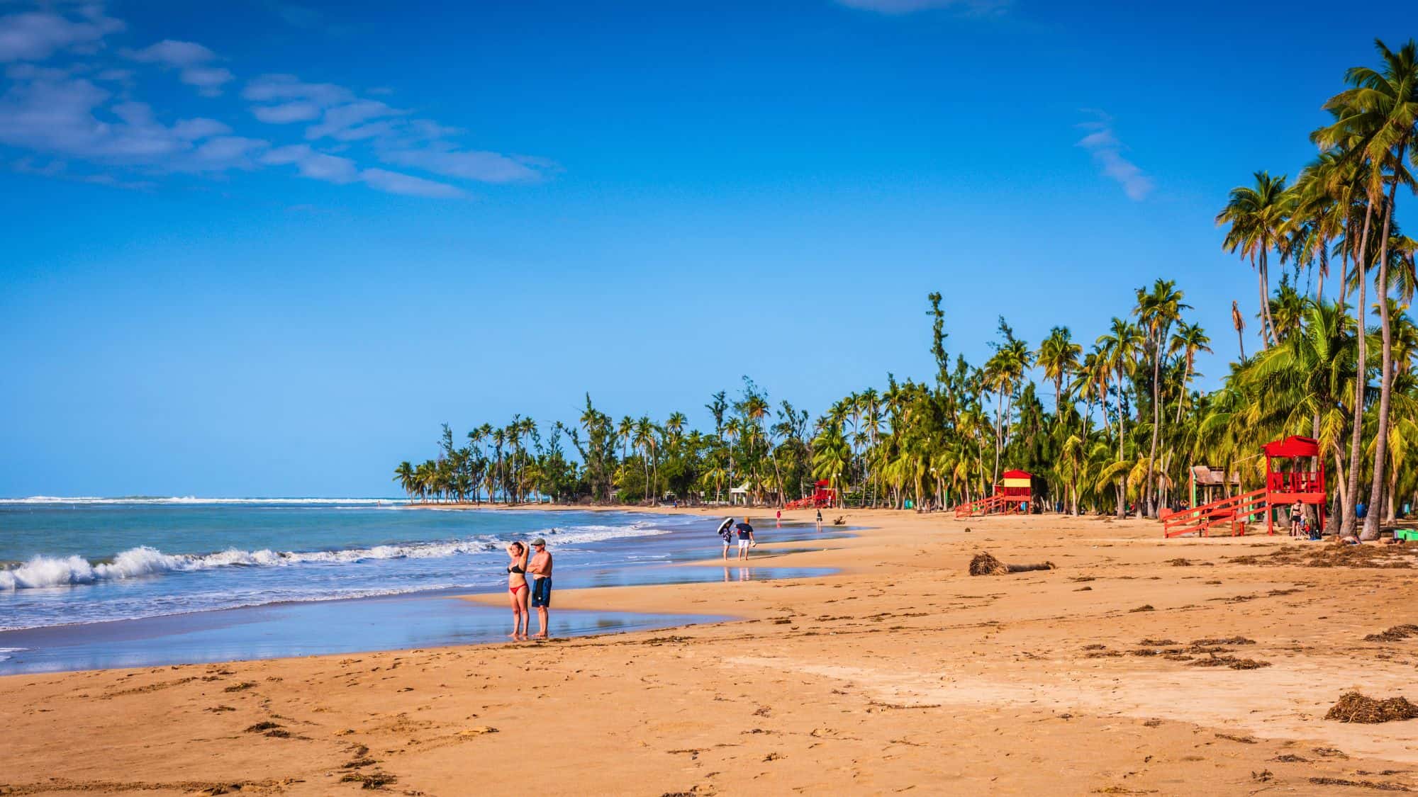 People stroll along the wide, sandy shoreline of Luquillo Beach, framed by tall palm trees and dotted with bright red lifeguard stands.