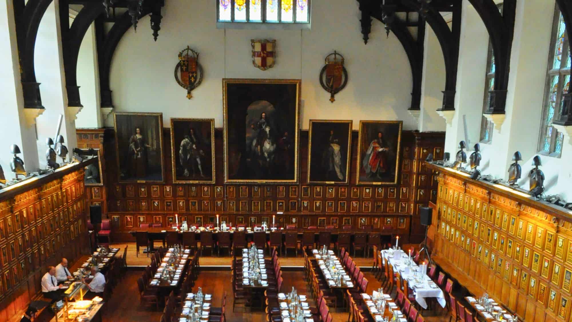 A grand dining hall with long wooden tables, medieval crests on paneled walls, and portraits of historical figures.
