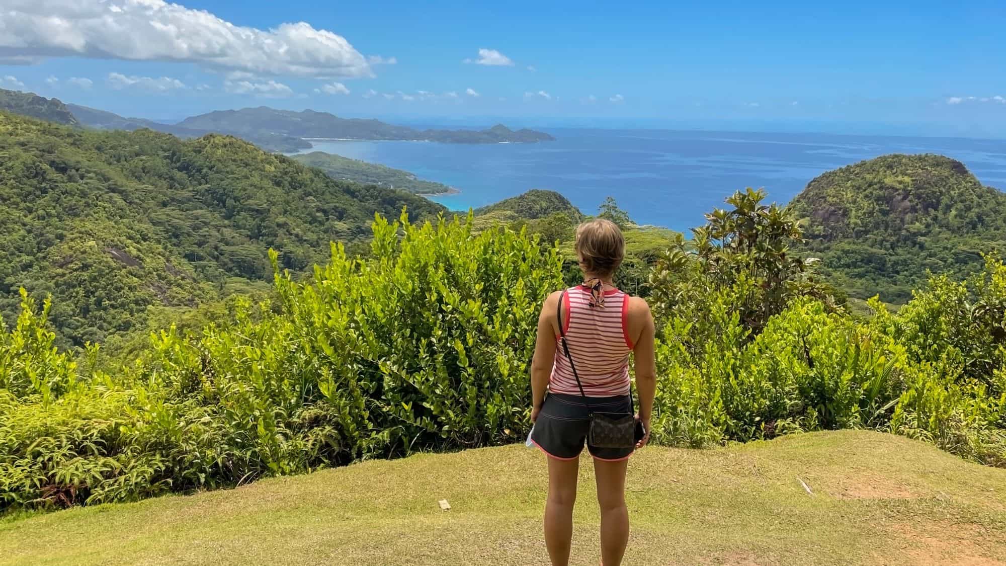 A woman in a striped tank top stands on a grassy overlook gazing at lush green hills and turquoise coastline.