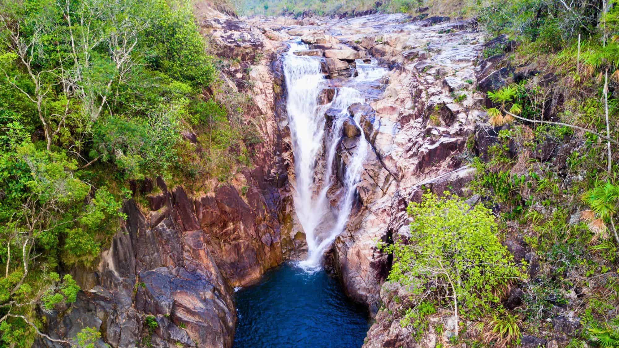 A tall waterfall cascading into a deep blue pool, framed by rugged cliffs and lush greenery.