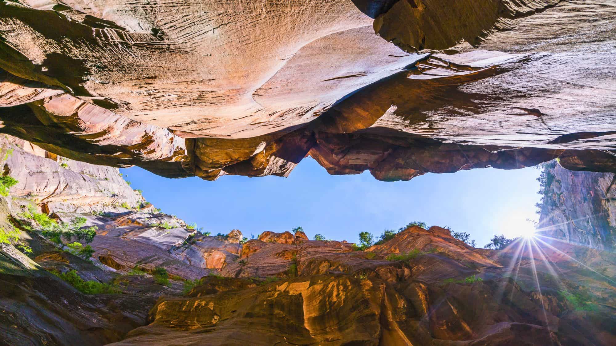 Looking straight up from the canyon floor, sheer rock walls frame a bright blue sky with the sun peeking through.