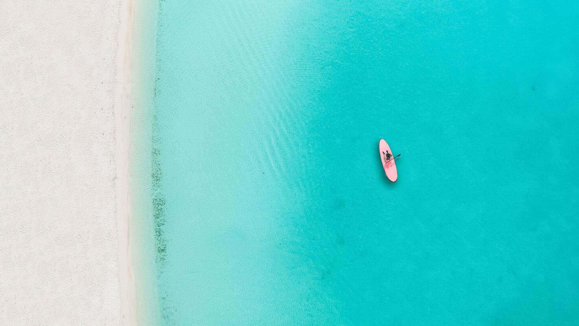 A person on a pink paddleboard floats over crystal-clear aqua water near a white sandy beach, viewed from above.