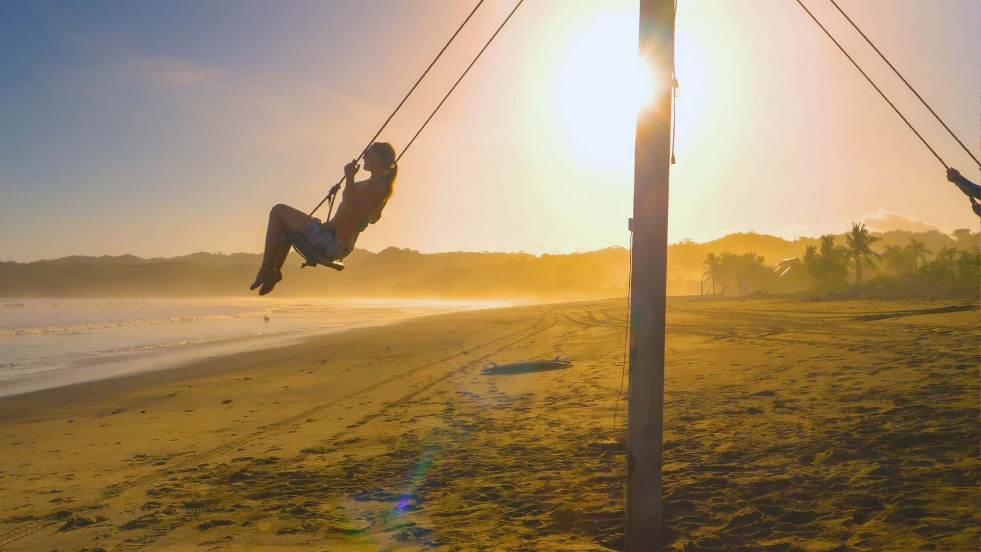 A woman swings high over a quiet sandy beach at sunset, with golden light casting long shadows and a surfboard resting nearby.
