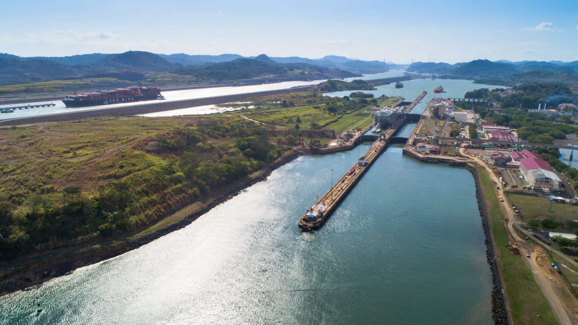 An aerial view of the Panama Canal shows massive cargo ships moving through the lock system, surrounded by industrial buildings and lush green hills.
