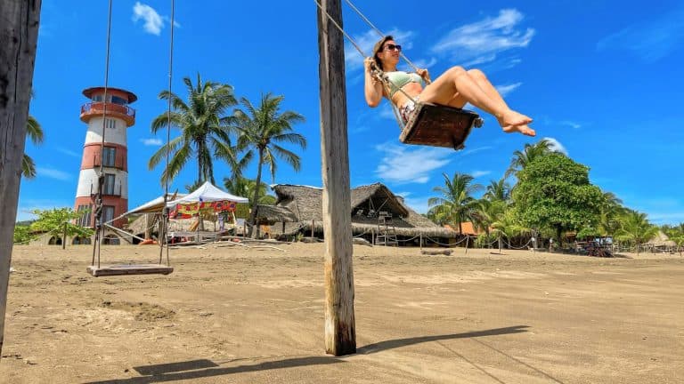 A woman smiles while swinging on a wooden swing set on a tropical beach, with a red-and-white striped lighthouse, palm trees, and thatched huts in the background under a vivid blue sky.