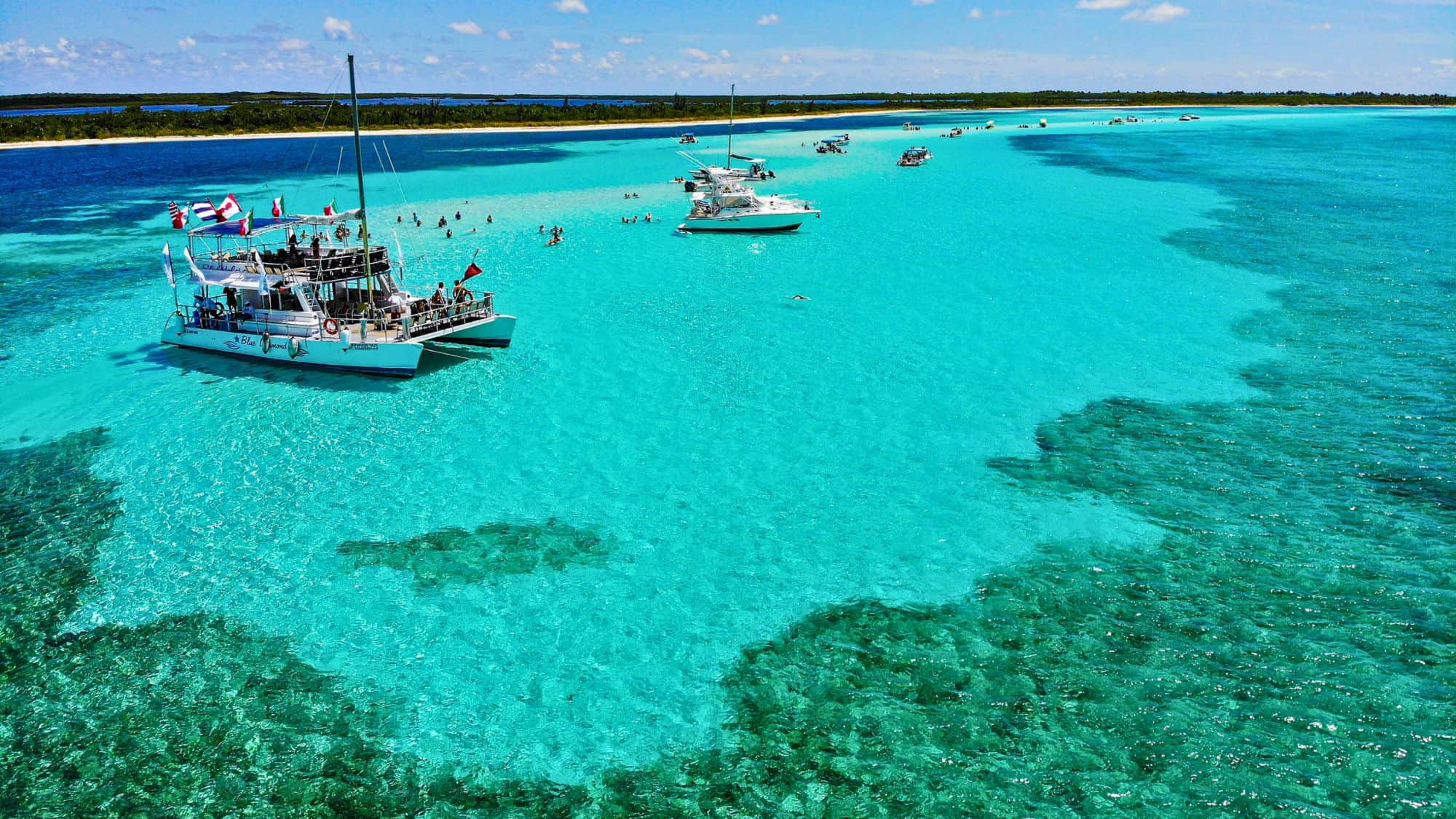 A large white catamaran floats in vivid turquoise water near a sandbar where people swim and wade.