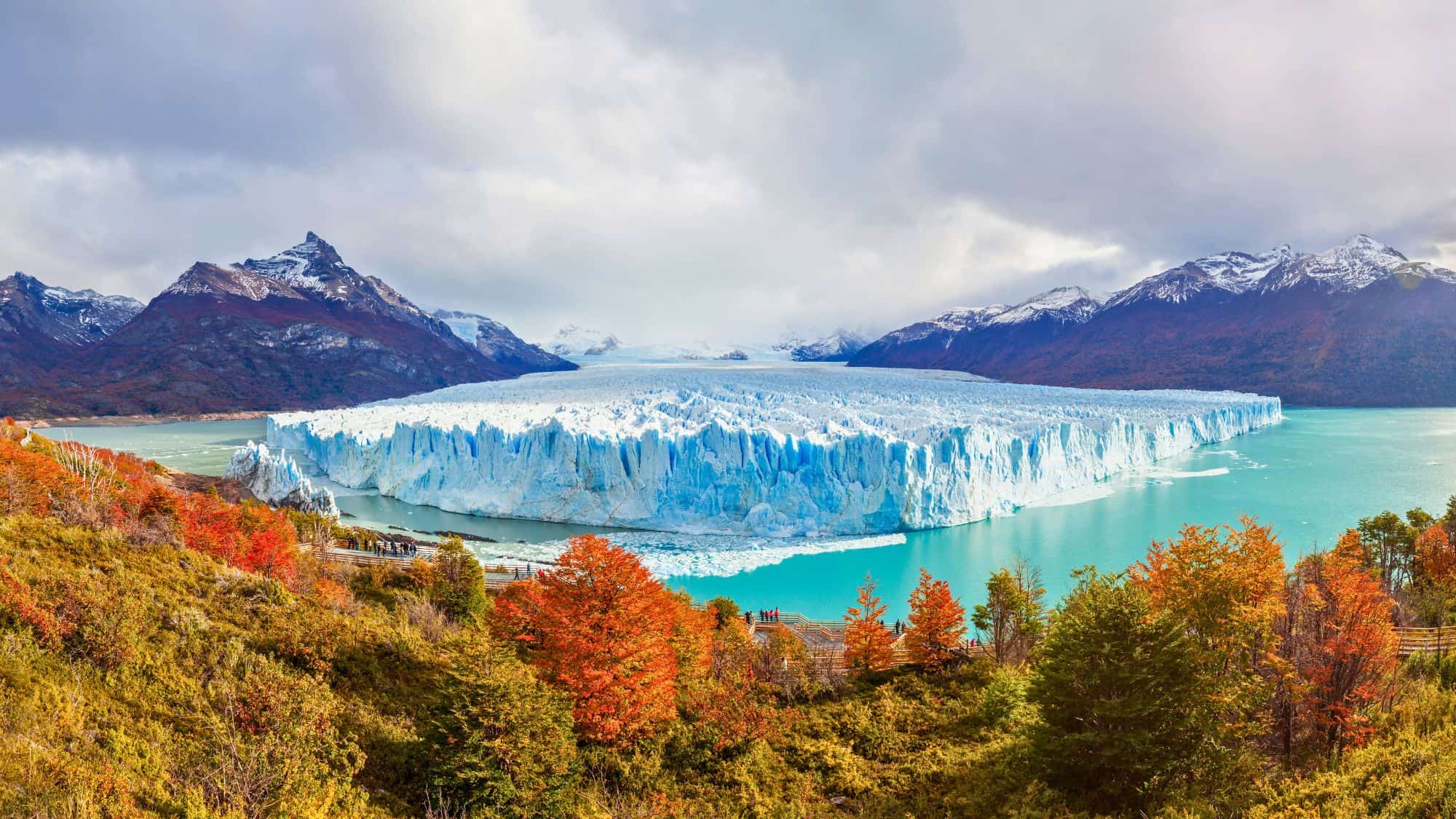 A massive blue glacier meets turquoise water, framed by colorful autumn trees and snow-dusted mountains.