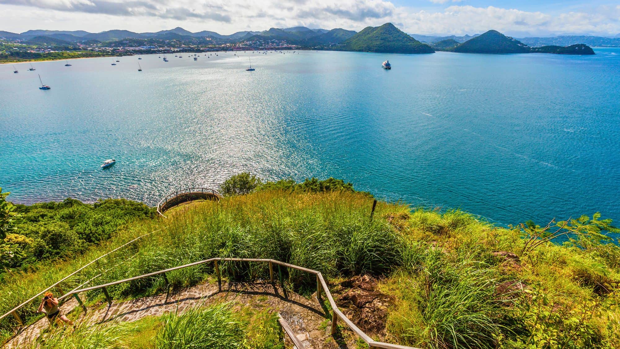 A winding staircase leads up a grassy hill overlooking a sparkling bay filled with boats and distant green peaks.