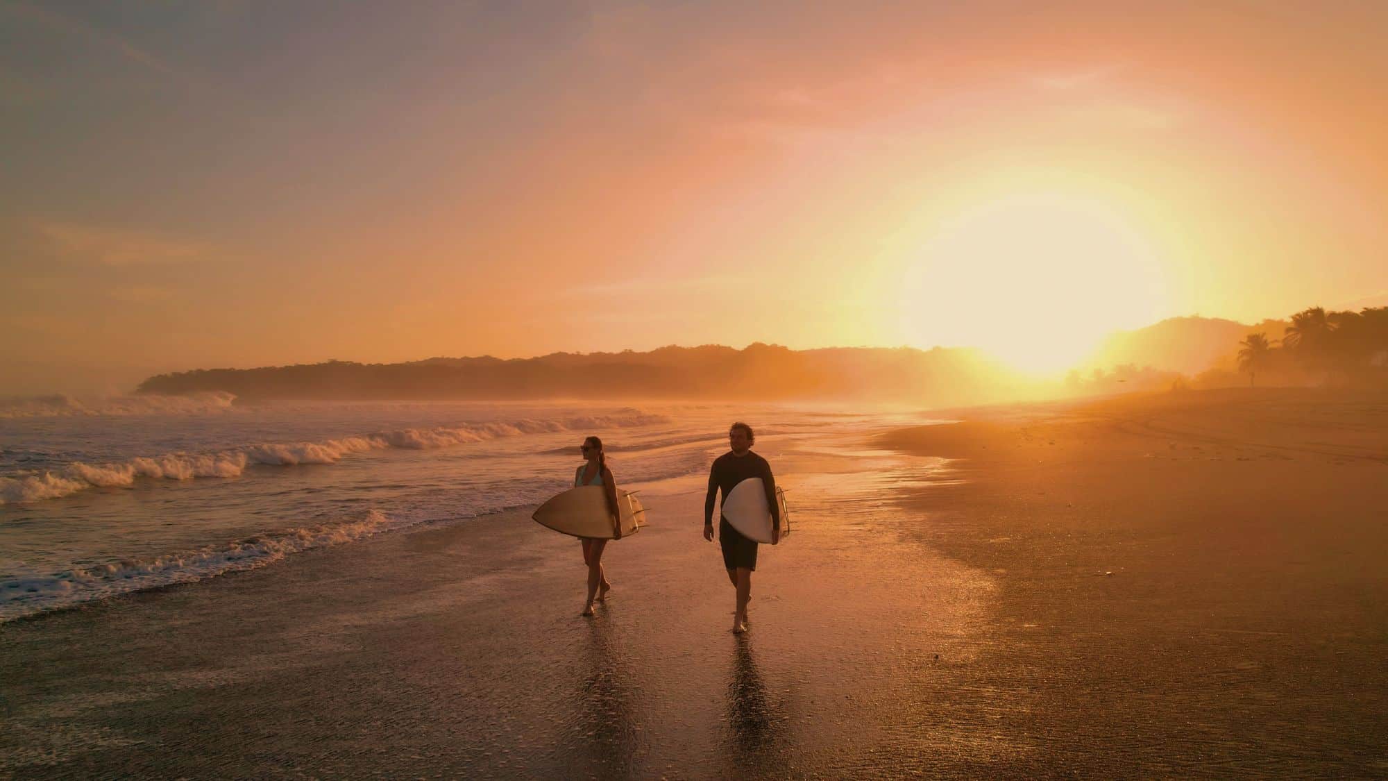 A man and woman carrying surfboards walk barefoot along a misty beach at sunset, the golden light reflecting off the wet sand and waves rolling beside them.