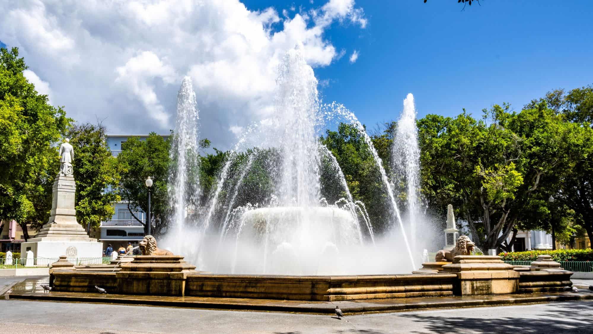 A large stone fountain with lion statues shoots water into the air at Plaza Las Delicias in Ponce, Puerto Rico, surrounded by trees and colonial architecture.