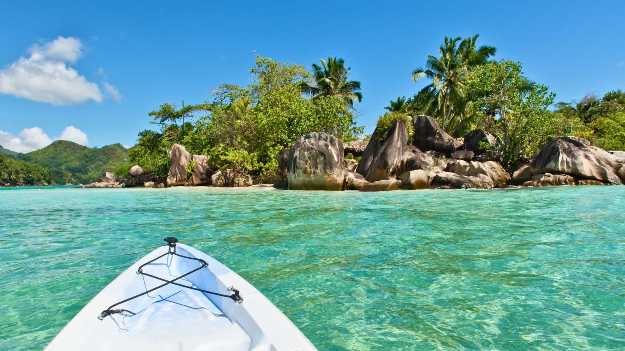 The bow of a kayak approaches a rocky, palm-fringed tropical shoreline over crystal-clear turquoise waters.