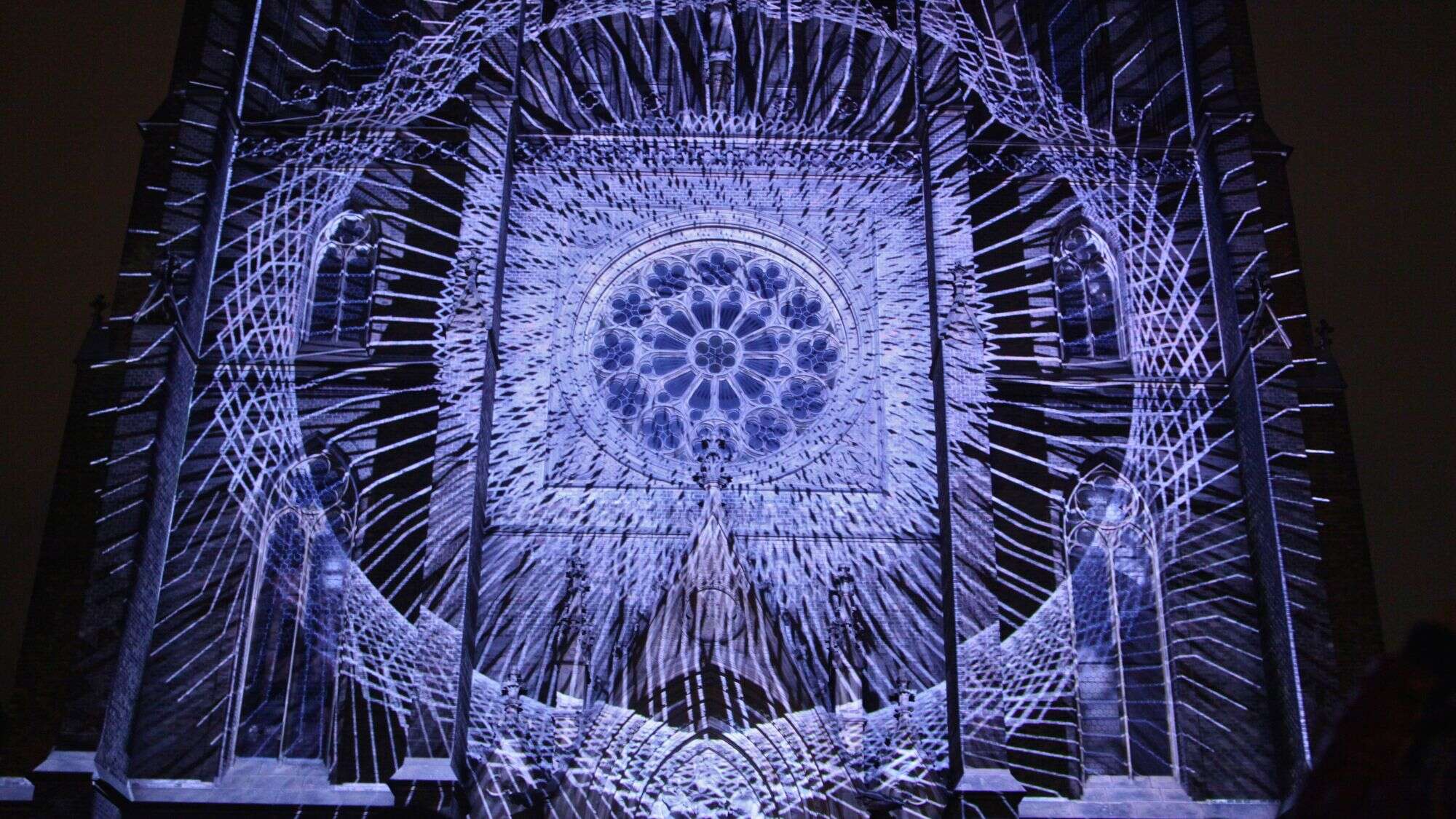 A cathedral facade is covered in intricate white light projections resembling a massive spiderweb or geometric pattern.