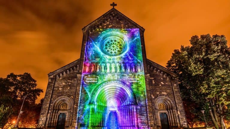 A stone church glows with rainbow-hued light projections against an orange night sky.