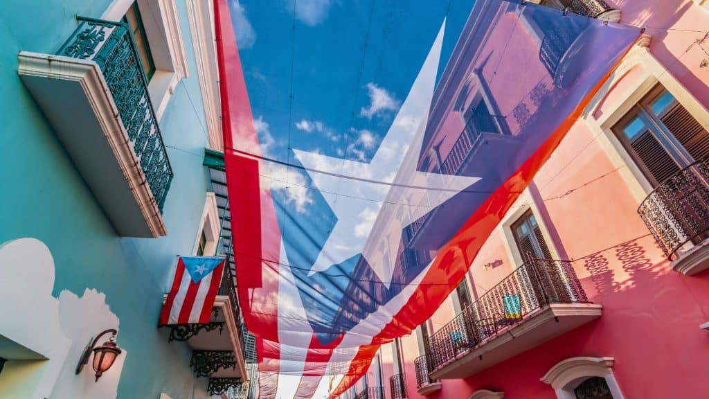A massive Puerto Rican flag canopy stretches across a narrow Old San Juan street lined with pastel buildings and balconies.