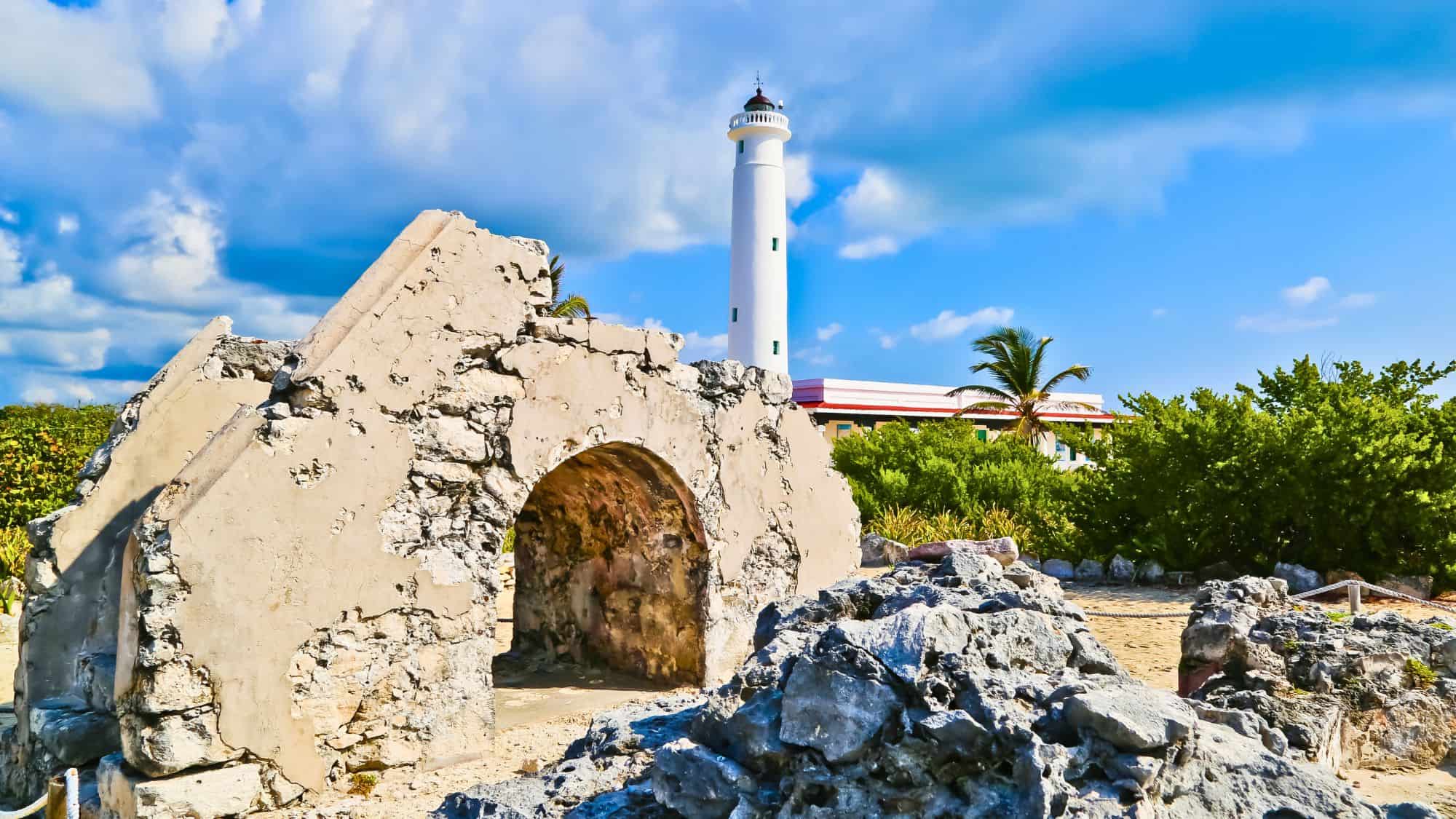 Crumbling stone ruins with an arched doorway stand in front of a tall white lighthouse under a partly cloudy sky.