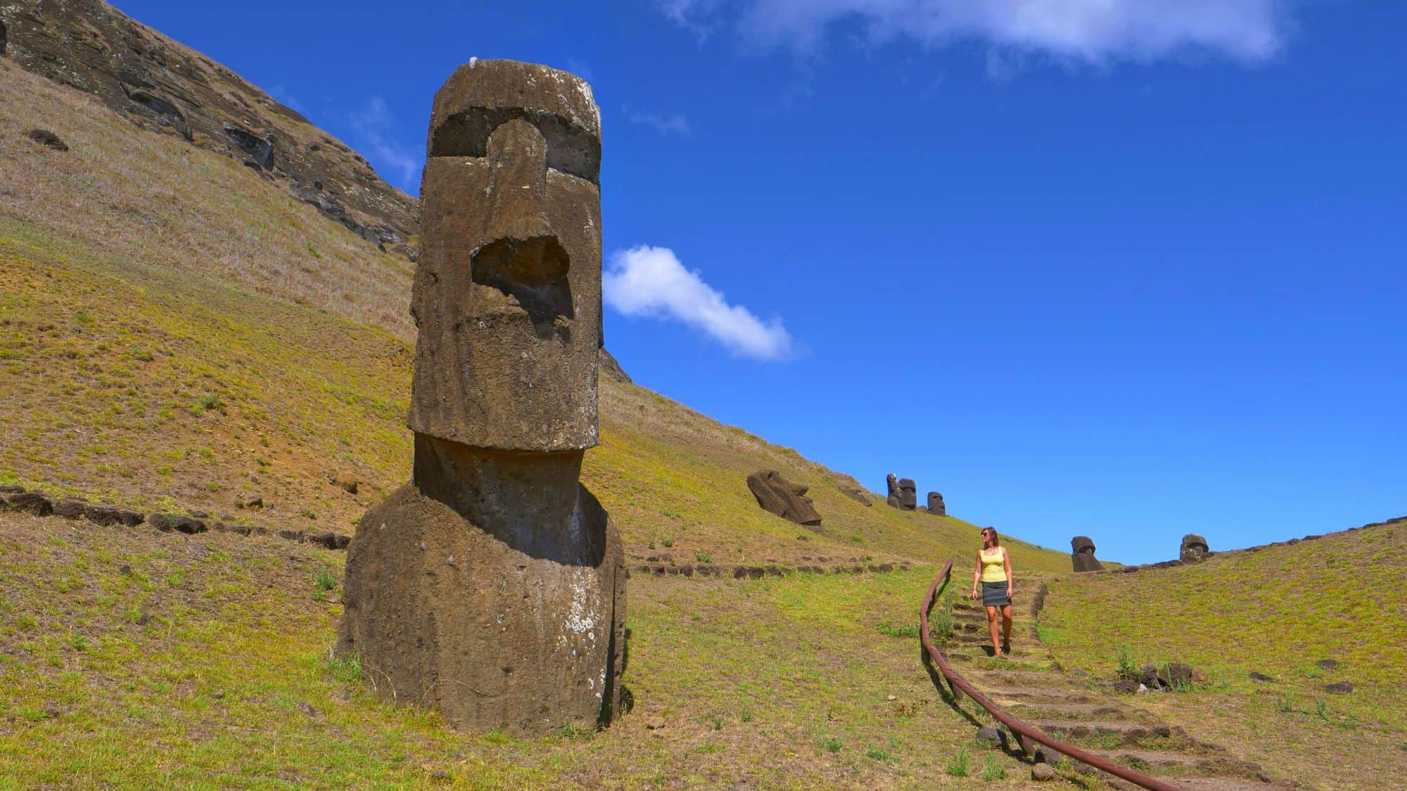 A woman walks down a hillside path past several partially buried moai statues on Easter Island.