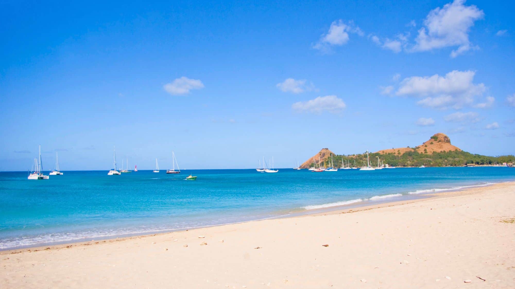 A wide sandy beach with calm turquoise waters and sailboats anchored offshore under a bright blue sky.