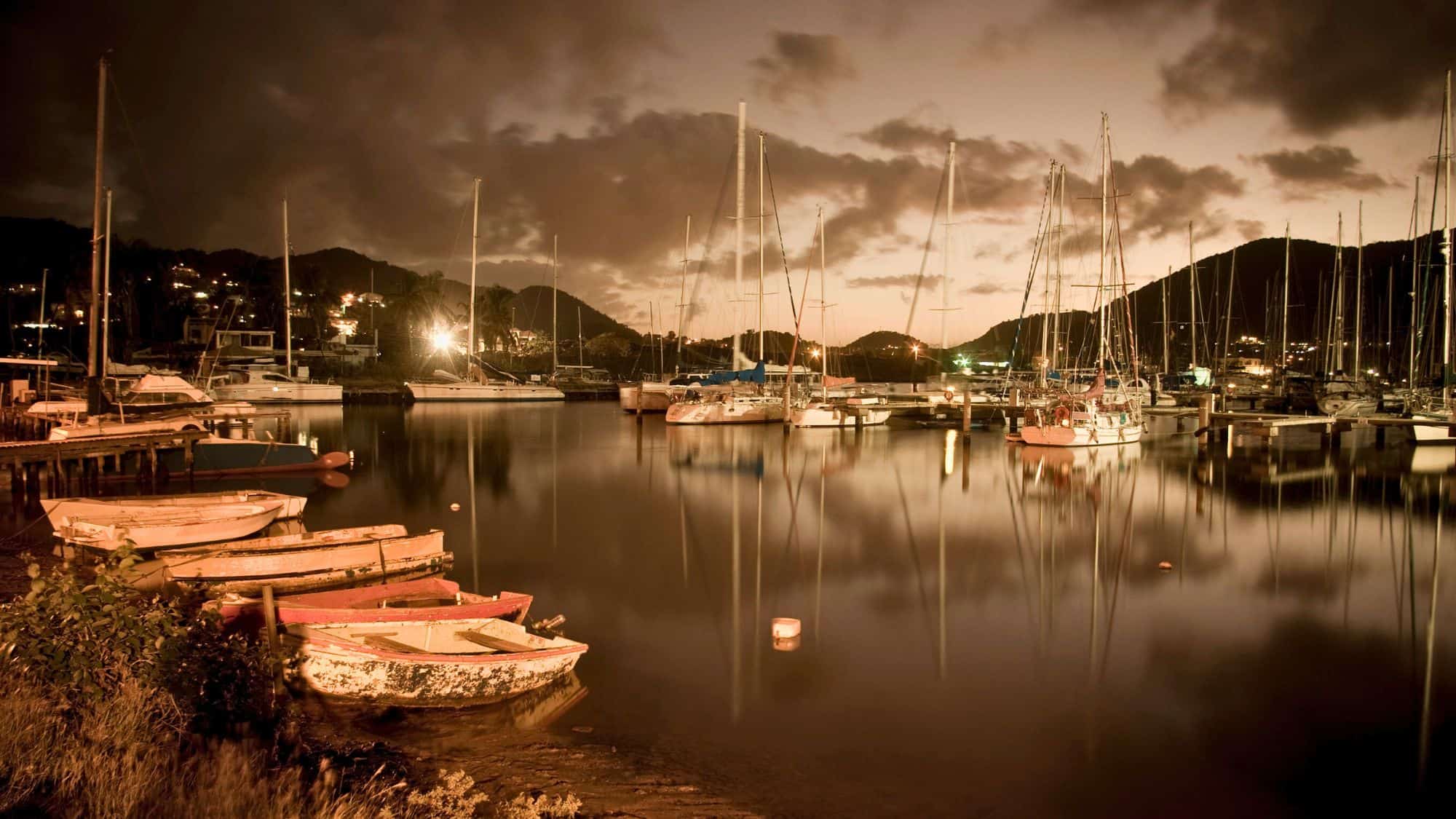 Wooden fishing boats and tall sailboats sit quietly in a dimly lit harbor, their reflections shimmering in the water at dusk.