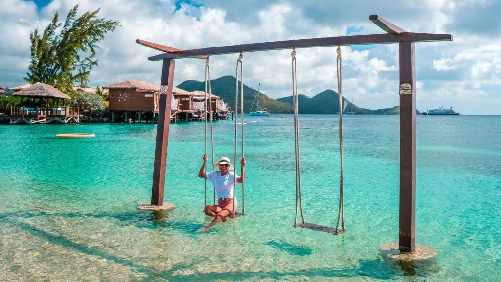 A person relaxes on a wooden swing set in shallow turquoise water near overwater bungalows and palm trees.