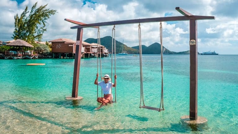A person relaxes on a wooden swing set in shallow turquoise water near overwater bungalows and palm trees.