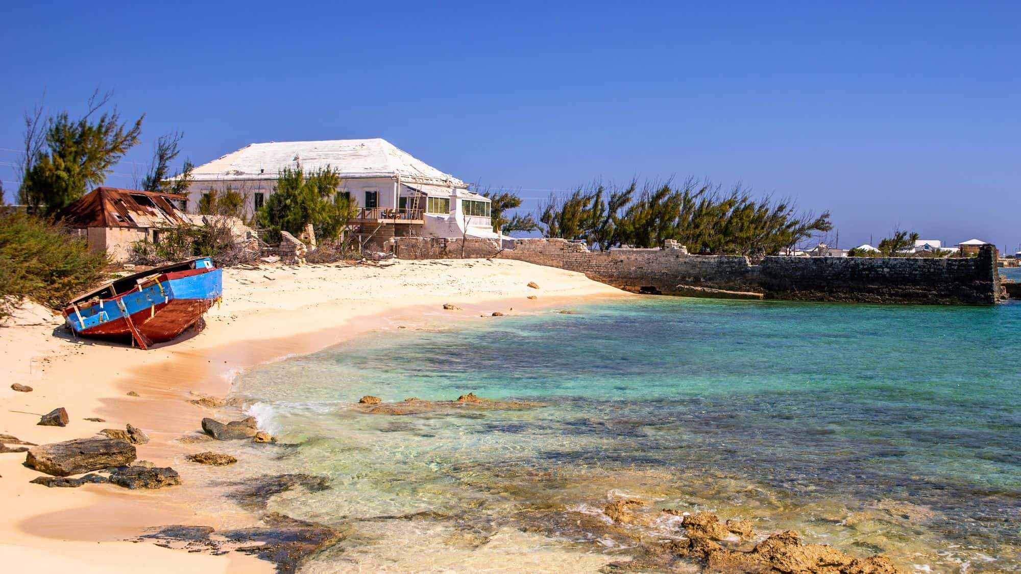 A weathered blue and red boat rests on a sandy beach near crumbling buildings and calm turquoise water.
