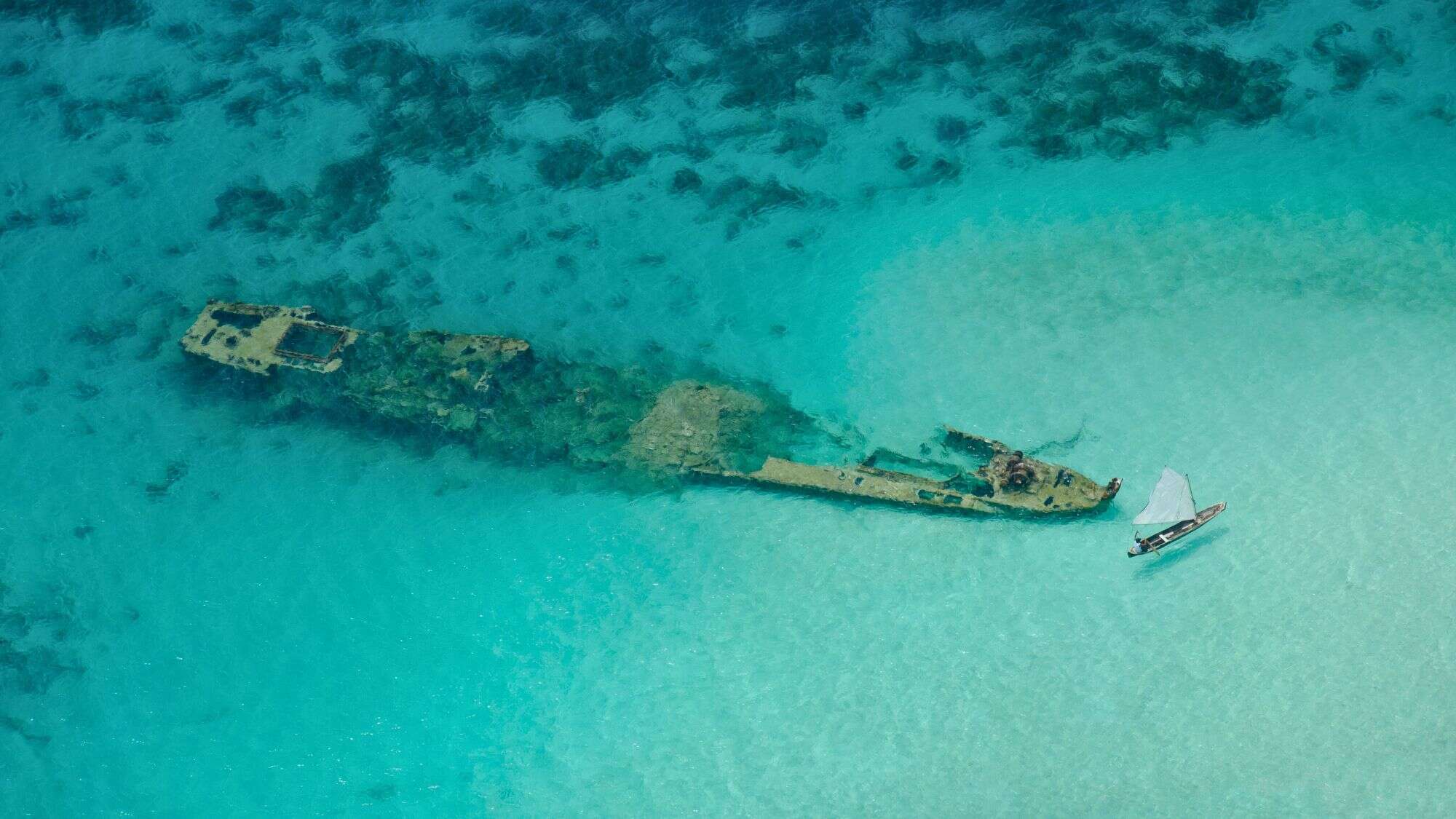 An aerial view of a rusted shipwreck partially submerged in shallow, crystal-clear turquoise water, with a small sailboat nearby emphasizing the scale and serenity.