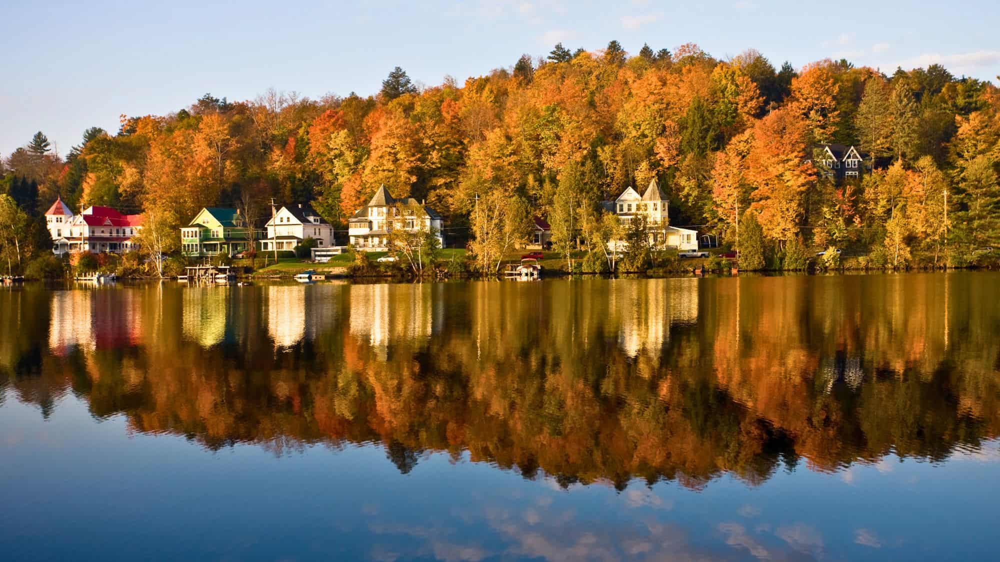 Victorian-style houses with colorful rooftops line the shore of Chautauqua Lake, reflected in still water backed by vibrant fall foliage.