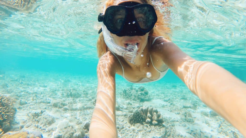 A woman takes an underwater selfie while snorkeling in clear turquoise water, with coral and rocks visible on the sandy ocean floor below.