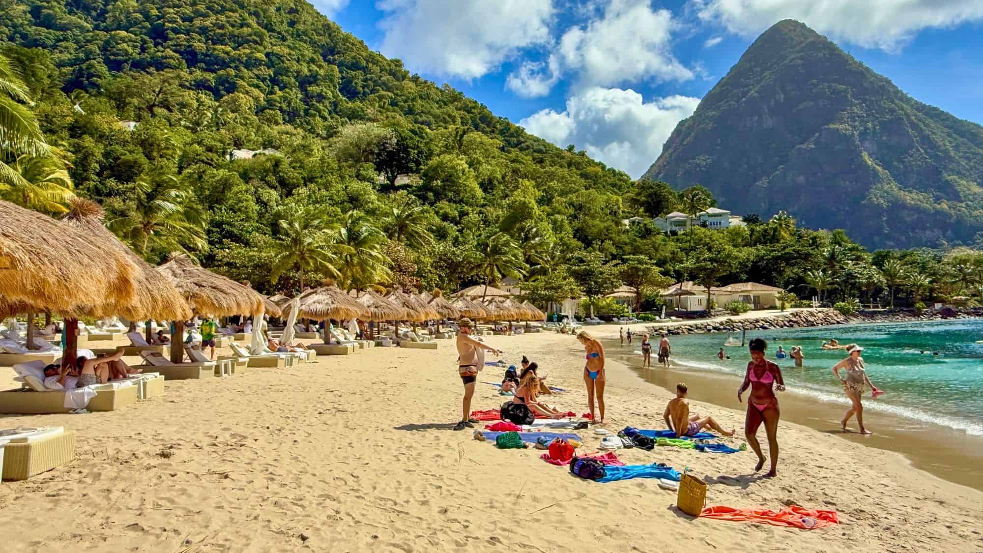 Tourists enjoy a sandy beach lined with thatched umbrellas and backed by the towering green Pitons.