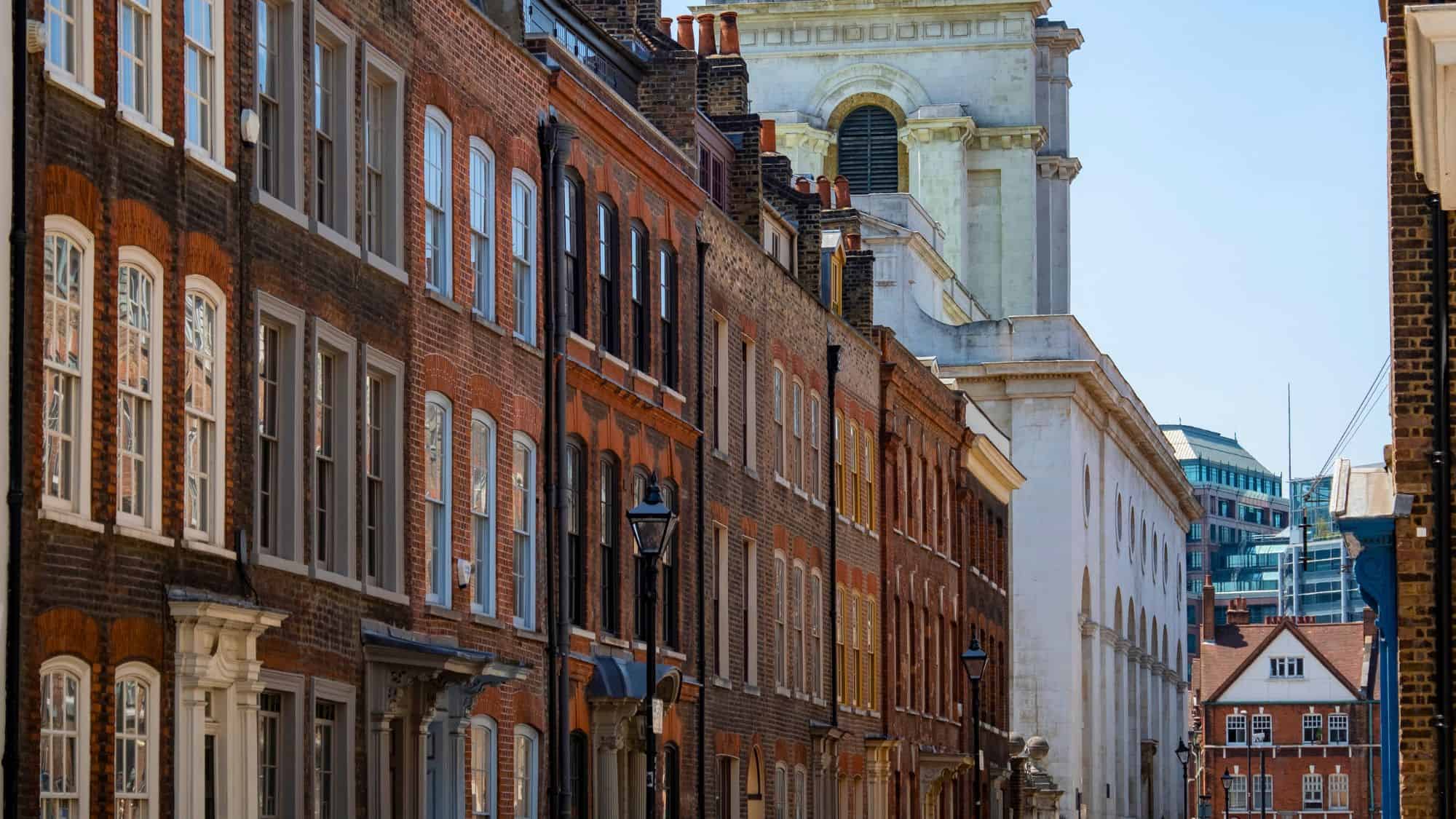A row of tall brick Georgian townhouses stands in the sunlight, with the white façade of a historic building towering behind.