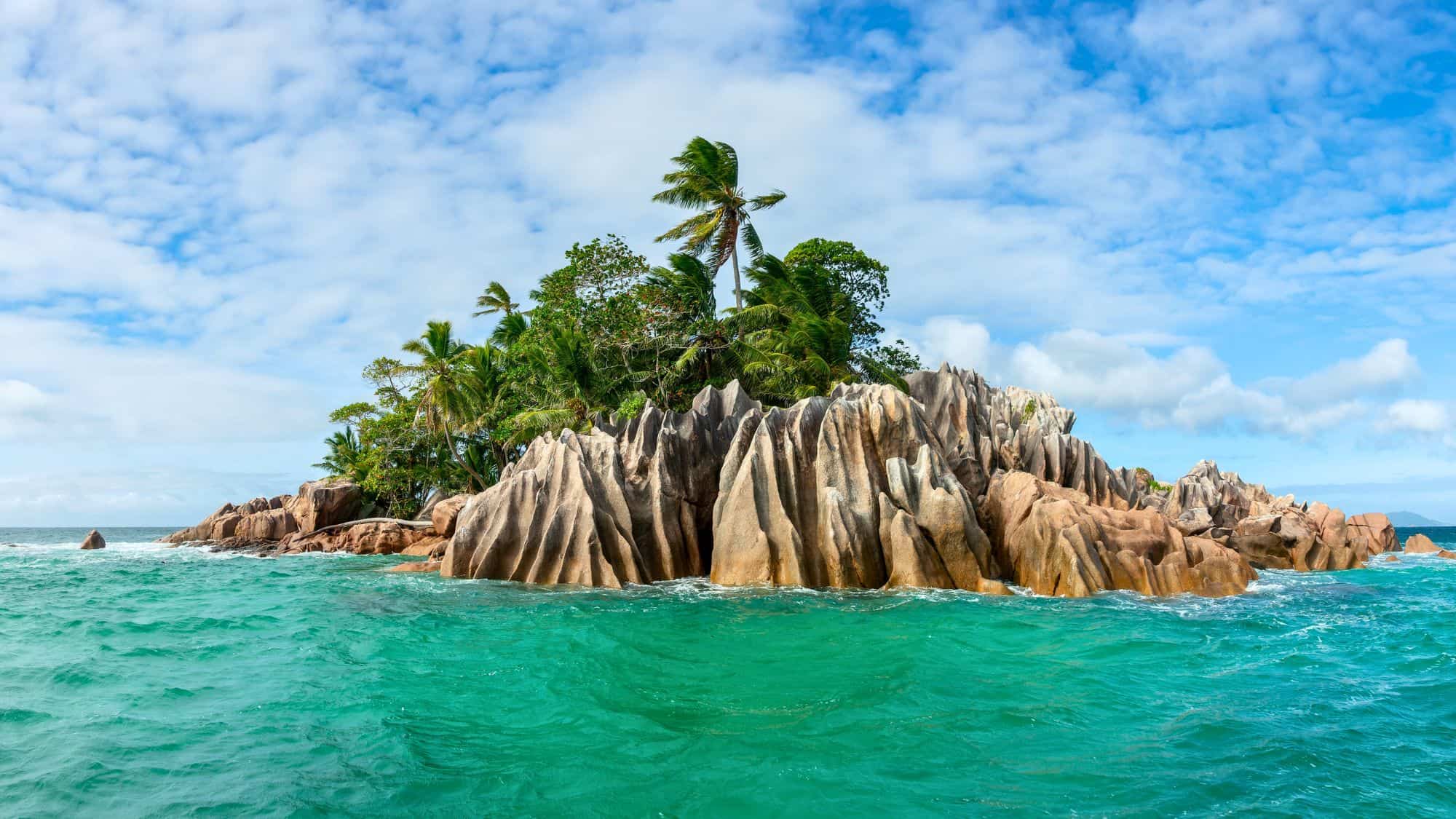 Jagged granite rock formations rise from a small palm-covered island surrounded by vibrant turquoise waters.