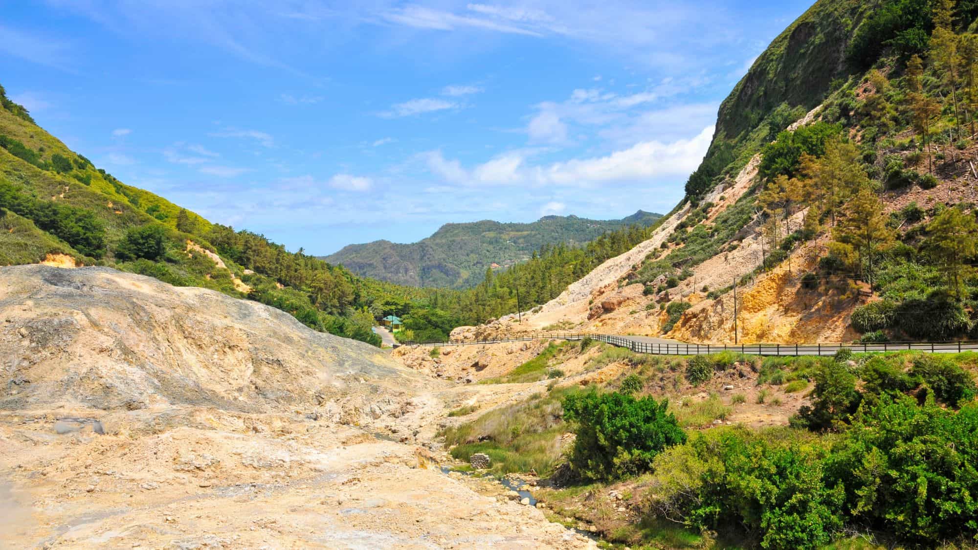 A geothermal landscape with steaming rocky terrain stretches between lush green mountains under a clear sky.
