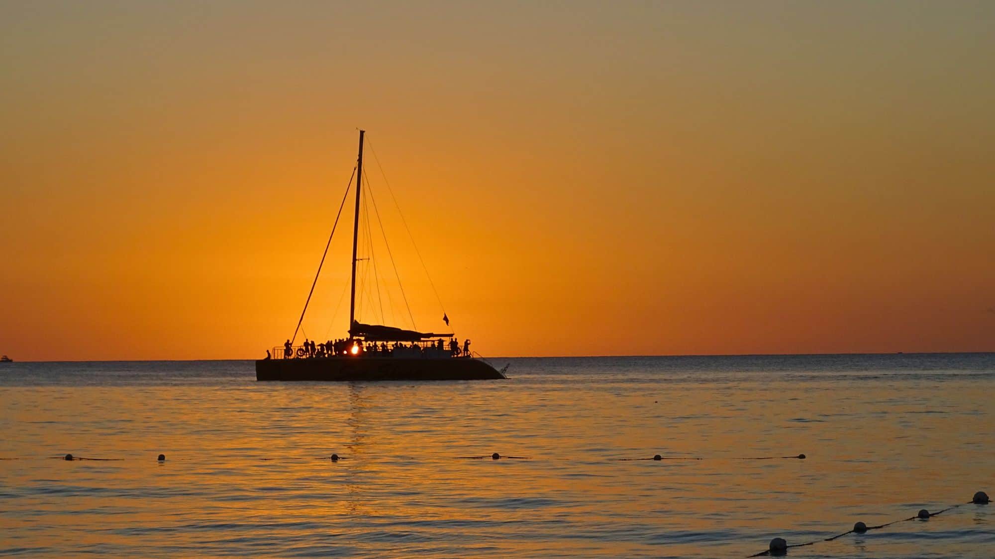 A catamaran full of passengers sails across calm waters at sunset, silhouetted against a fiery orange sky.