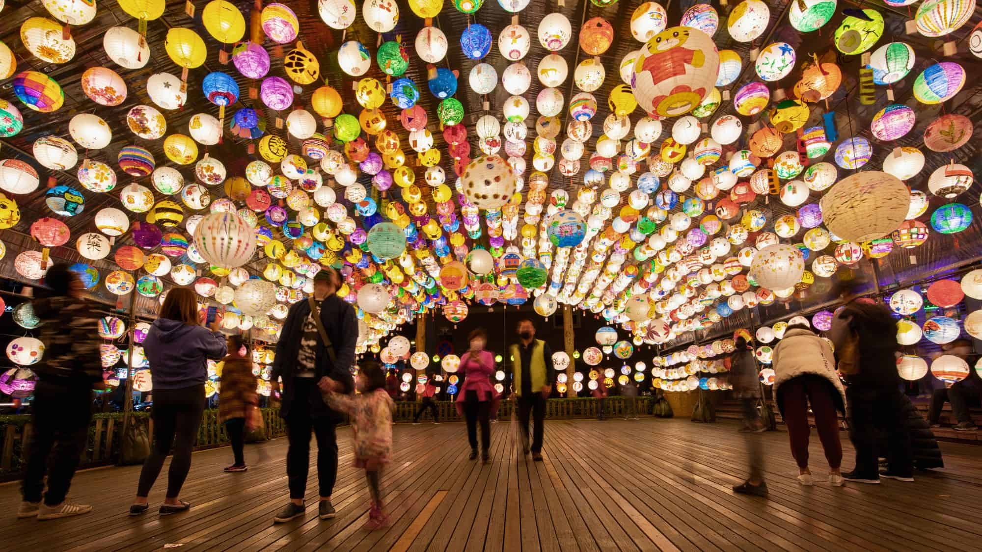 People stroll under an arched ceiling of hundreds of colorful, hand-painted lanterns at a nighttime festival.