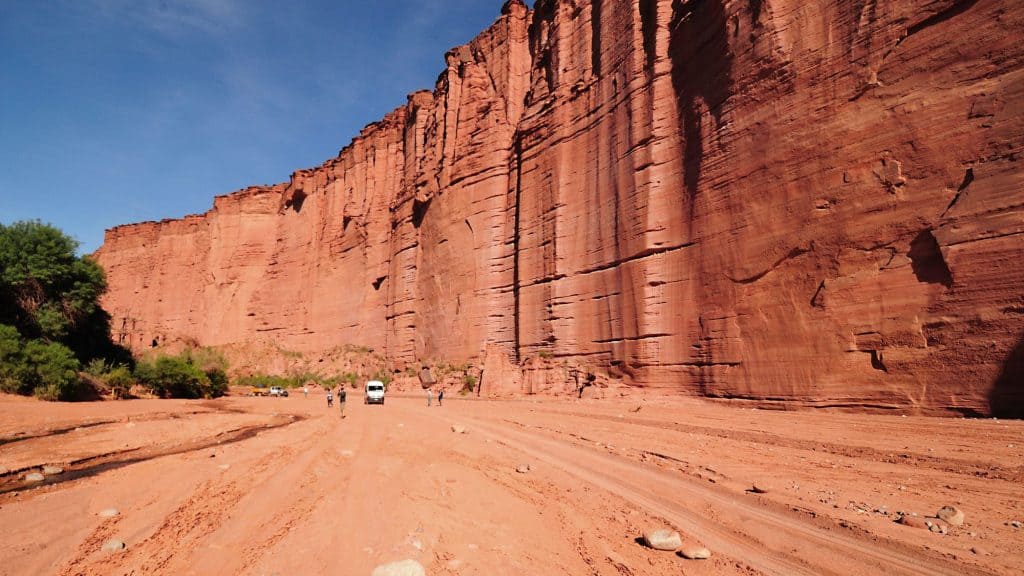 Sheer red sandstone cliffs tower over a wide sandy trail where people and vehicles appear small below.