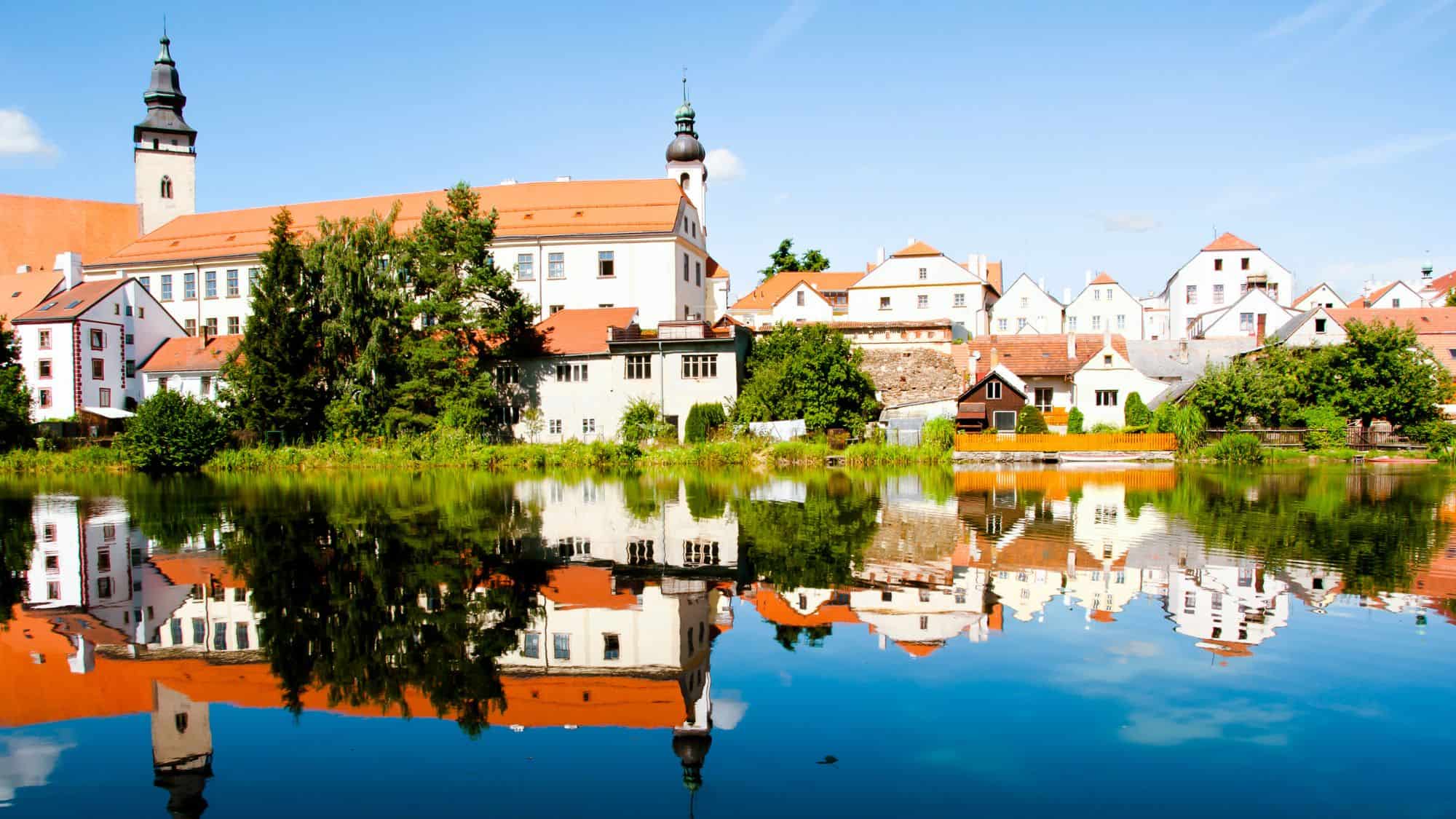 The historic town of Telč is mirrored in the calm surface of a pond, with whitewashed houses and church spires.