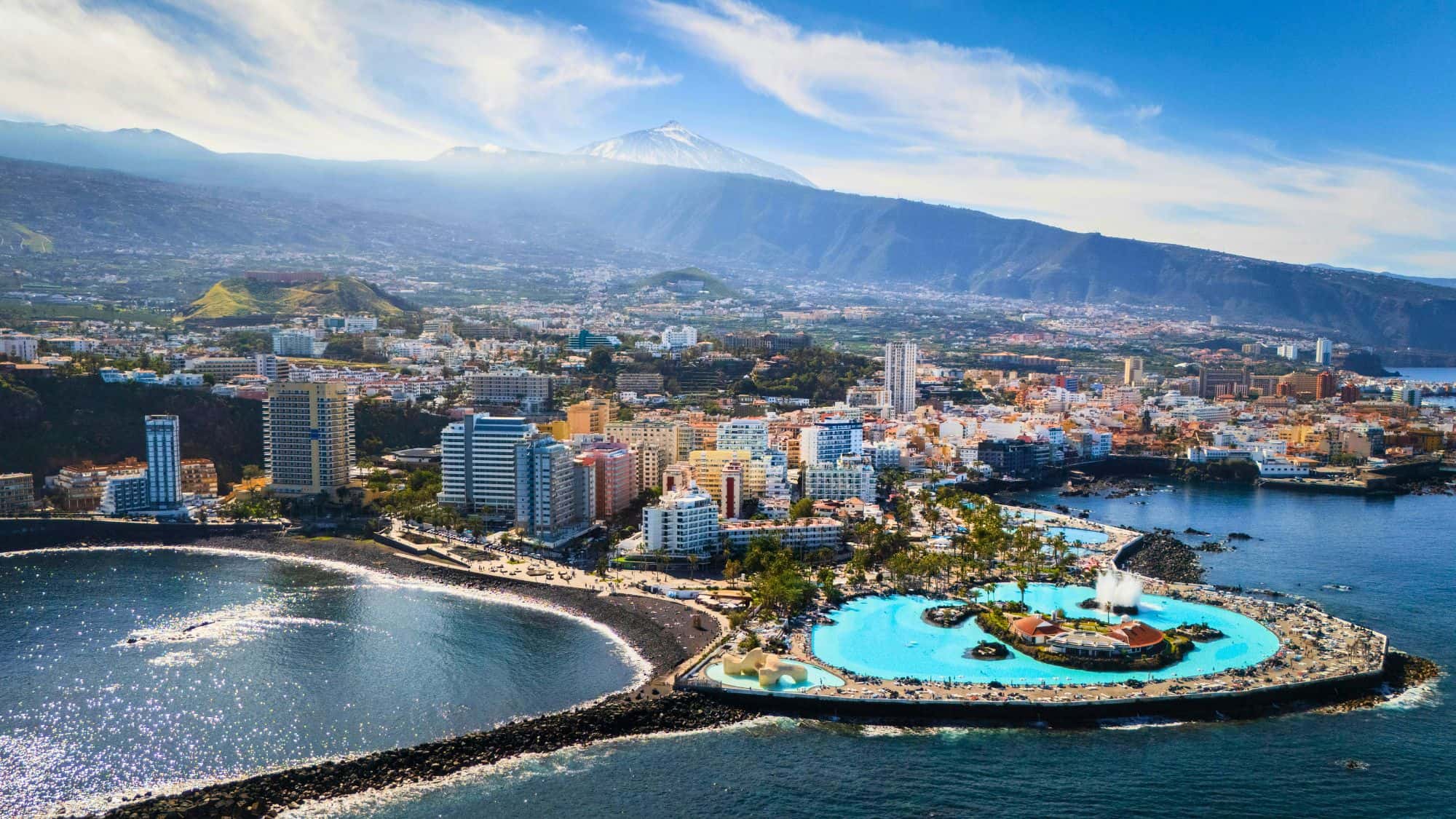 An aerial view of Tenerife shows a vibrant city, sandy beaches, and a massive seaside pool with Mount Teide in the distance.