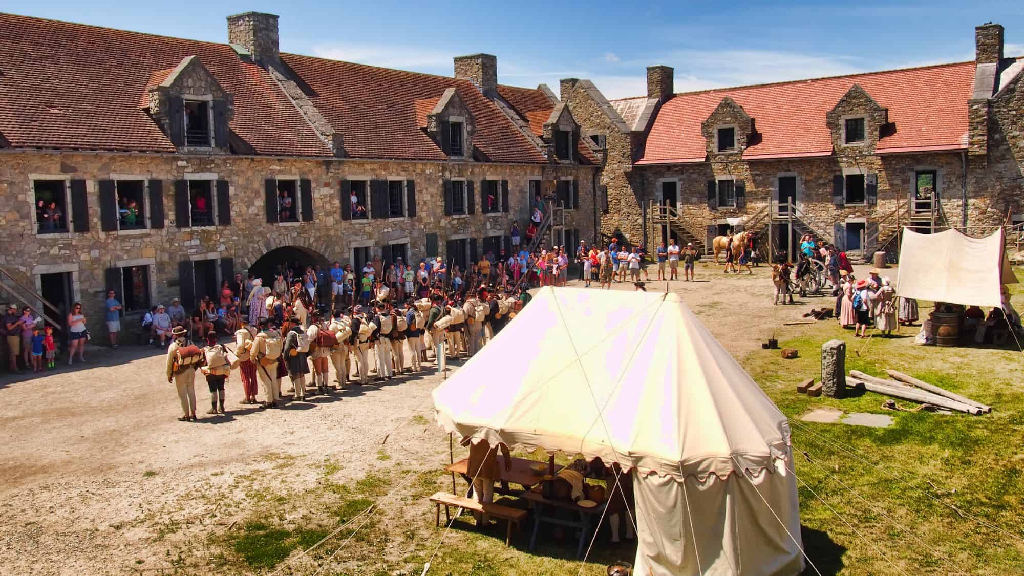 Historical reenactors in period dress assemble in the courtyard of Fort Ticonderoga, surrounded by stone buildings and tents with a crowd of visitors watching.