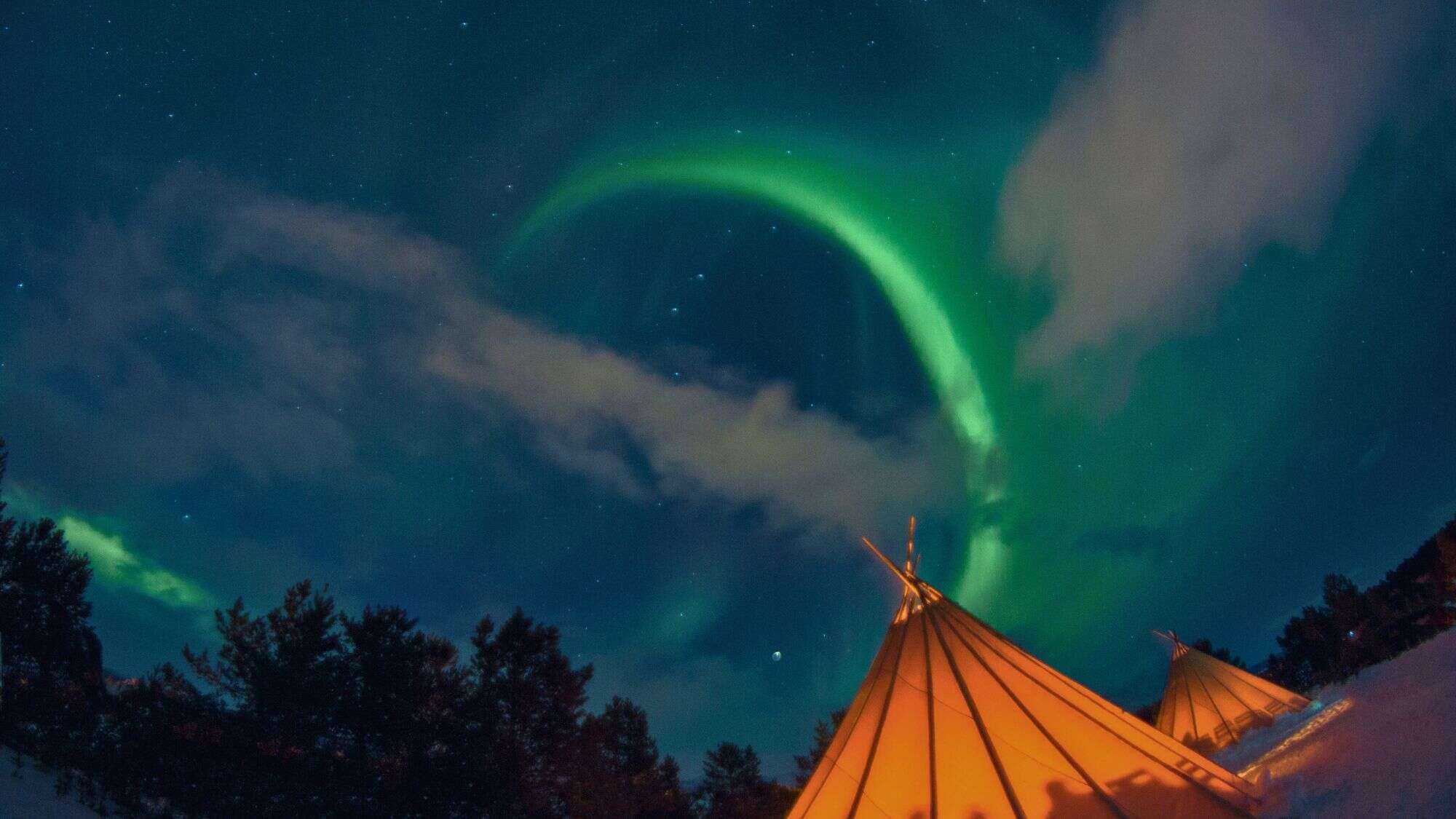 A green aurora arcs across the starry sky above glowing orange teepees in a snowy forest.