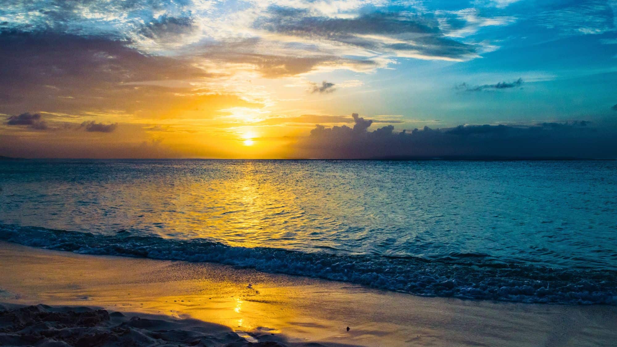 The sun sets over the horizon, casting golden reflections across the calm ocean waves and wet sand under a dramatic, cloud-streaked sky.